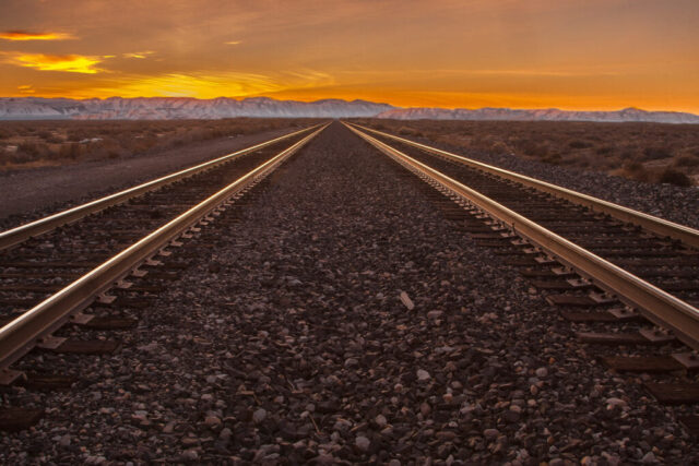 Golden desert sunset photograph captured along the twin railroad tracks near Flanigan, Nevada — a remote stretch of high desert once home to a bustling rail junction. The glowing rails lead the viewer’s eye toward the horizon, where the last light of day ignites the clouds above the distant Smoke Creek Mountains. Once an important crossroads for the Western Pacific and Nevada-California-Oregon railroads, Flanigan now stands quiet, its history fading into the open expanse of sagebrush and sky. This image embodies the solitude and timeless beauty of the American West — a place where steel, dust, and sunset meet at the vanishing point.