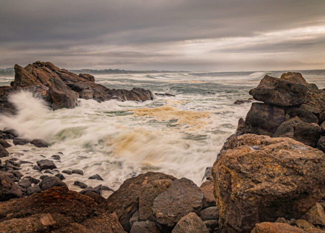 Powerful Pacific waves crash against dark basalt rocks along the rugged shoreline of Yaquina Head near Newport, Oregon, under a brooding autumn sky. The scene captures the raw energy of the Oregon Coast, with swirling sea foam and mist rising from the churning surf beneath layered gray clouds. Formed by ancient lava flows, the jagged coastline showcases the dramatic geology that defines this iconic headland, home to the historic Yaquina Head Lighthouse. This coastal landscape photograph evokes the untamed beauty and moody atmosphere of the Pacific Northwest, ideal for lovers of seascape photography, Oregon’s wild shores, and the natural drama of the Yaquina Head Outstanding Natural Area.