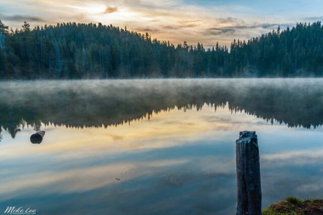 Peaceful dawn over Echo Lake in Lassen National Forest, Northern California, captured on a calm late May morning. Soft golden sunlight breaks through streaks of cloud above a mirror-still lake, where faint mist drifts across the water and evergreen pines reflect perfectly along the shoreline. A weathered wooden post in the foreground adds depth and texture to the tranquil composition. This fine art landscape photograph evokes the quiet beauty of nature at sunrise, celebrating a serene wilderness moment before a day of fishing and father-son adventure.