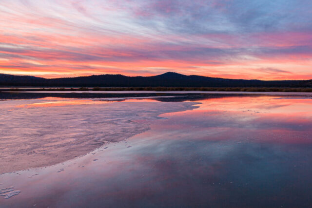 Icy Sunset Peaceful winter sunset photograph captured on the north shore of Eagle Lake near the community of Spalding in Lassen County, Northern California. The icy lake reflects soft pastel hues of pink, orange, and lavender as the sun dips below the horizon, casting a gentle glow across the frozen shallows. In the distance, dark hills and brush frame the tranquil expanse of water once known to anglers as “Heart Failure,” a name as rugged as the road leading there. This fine art landscape conveys the quiet beauty of a crisp January evening — where calm skies, glassy ice, and fading light meet in perfect balance on one of Northern California’s most scenic alpine lakes.