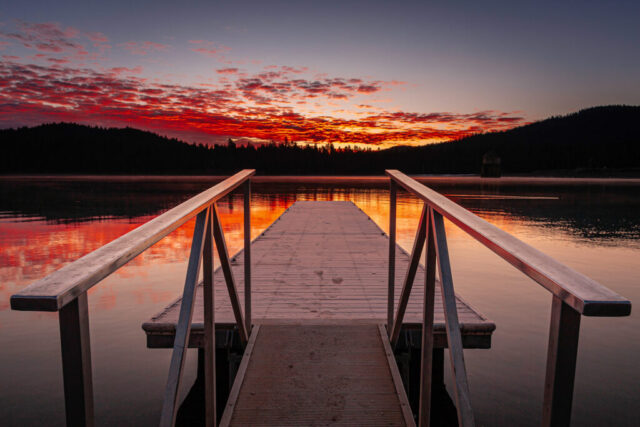 Almanor Dawn Stunning sunrise photograph taken from the dock at Canyon Dam boat launch on Lake Almanor in Plumas County, Northern California. The warm glow of dawn ignites the clouds in brilliant shades of red, orange, and gold, reflecting perfectly across the glassy lake surface. The frost-dusted dock leads the viewer’s eye toward the horizon, where the silhouetted tree line meets the fiery morning sky. Lake Almanor, a vital Northern California reservoir holding 1.3 million acre-feet of water, is not only a key hydroelectric source but also a beloved year-round recreation destination. This tranquil scene captures the peaceful beauty and reflective stillness of a Sierra sunrise.