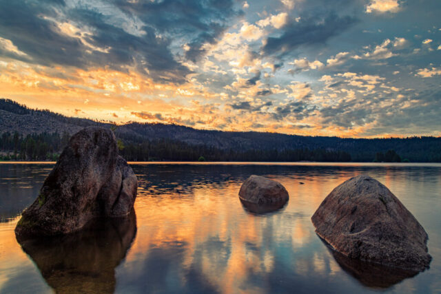 Peaceful sunrise photograph over Antelope Lake in Plumas County, Northern California, captured on a calm late May morning. The early light spills across the lake’s smooth surface, illuminating scattered boulders in the shallows and reflecting the brilliant golden and blue hues of the sky above. Layers of forested ridgelines frame the background, while textured clouds add dramatic depth to the dawn. Surrounded by quiet wilderness and rich in wildlife, Antelope Lake offers a serene glimpse of the Sierra Nevada at its most tranquil — a moment of still beauty where water, sky, and land come together in harmony.