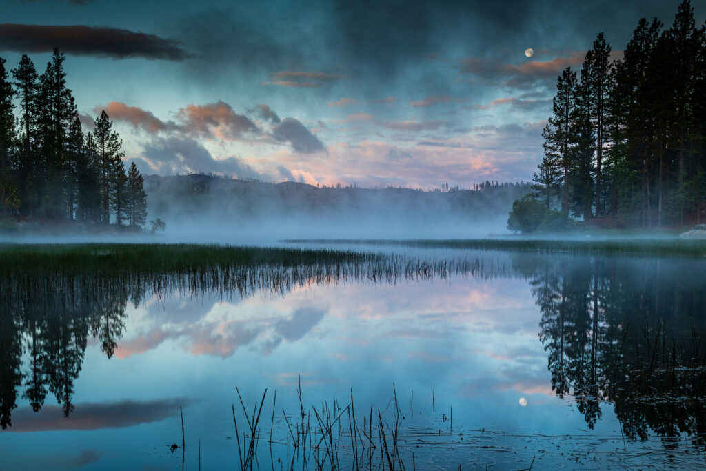 Dreamlike blue-hour scene at Antelope Lake in Plumas County, California, captured as mist rises from the warm water after an early June storm. The setting Strawberry Moon glows above layers of dramatic clouds and calm reflections, framed by tall pine silhouettes and soft meadow grasses. The photograph blends serenity and power, revealing the atmospheric beauty created by cold air meeting the lake’s surface warmth. This award-winning landscape image from 2020 embodies the peaceful allure of the Sierra Nevada wilderness and the tranquil magic of dawn over Northern California’s mountain lakes.