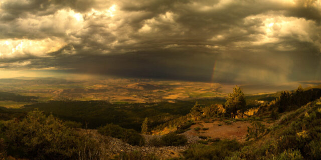 Panoramic landscape photograph titled “Passing Storm,” captured from near the summit of Thompson Peak on the Lassen–Plumas County Line in Northern California. The 2:1 composition overlooks Honey Lake Valley in Lassen County, where sunlight breaks through a retreating summer thunderstorm at sunset. Dramatic clouds tower above the vast valley floor, streaked with rain and a faint rainbow, while the golden light casts warm tones over the rugged terrain, pine-covered slopes, and distant hills of Bald Mountain and Bass Hill. The scene captures the fleeting beauty of storm light over Janesville, California, blending natural drama and tranquility in a fine art landscape photograph.