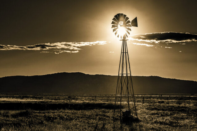 Old Reliable Sepia-toned photograph of a classic ranch windmill silhouetted against the setting sun near Madeline, California. The sun glows through the windmill blades, casting long rays across a vast open plain bordered by low hills and a streak of distant clouds. The warm monochrome treatment enhances the nostalgic feel, evoking the timeless spirit of rural life and the enduring presence of wind pumps across the Western landscape.