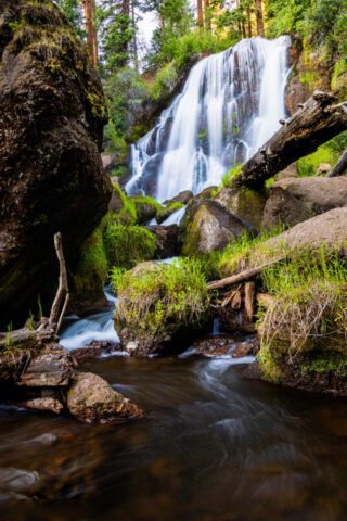 Mill Creek Falls Vibrant color photograph of Mill Creek Falls near Likely, in remote Modoc County, Northern California. Captured from the base of the falls in late afternoon light, the image highlights the graceful flow of water cascading over moss-covered rocks surrounded by lush green foliage and tall pines. The soft, silky effect of the long exposure contrasts beautifully with the rugged textures of volcanic stone and fallen timber, creating a dynamic yet tranquil composition. This fine art nature image captures the untouched beauty of the South Warner Wilderness and is perfect for cabin décor, mountain homes, or anyone who loves serene waterfall landscapes and the wild charm of Modoc County.