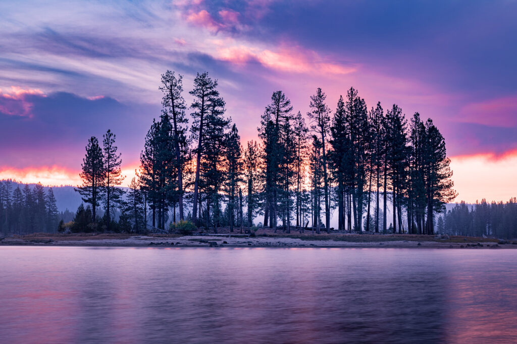Peaceful sunrise landscape photograph of Dedication Island at Antelope Lake in Plumas County, Northern California. The first light of dawn filters through a stand of tall pines, painting the sky in soft hues of violet, pink, and gold. Gentle mist rises from the lake’s calm surface, adding a quiet stillness to the scene. The silhouettes of the trees stand sharply against the radiant morning sky, reflecting the timeless beauty of this remote Sierra Nevada reservoir. This fine art image captures the serene moment when night gives way to day in one of Northern California’s most tranquil mountain settings.