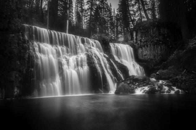 Dreamy Cascade Black and white photograph of Middle McCloud Falls in Northern California, captured on a crisp autumn morning at 28°F. The long exposure transforms the powerful waterfall into a soft, dreamlike cascade flowing over dark basalt rock, framed by tall evergreens that fade gently into the misty dawn light. The stillness of the empty scene conveys solitude and serenity, emphasizing the natural grace and strength of the falls. This fine art landscape image embodies the quiet magic of early morning in the wilderness, ideal for nature lovers and admirers of tranquil monochrome photography.