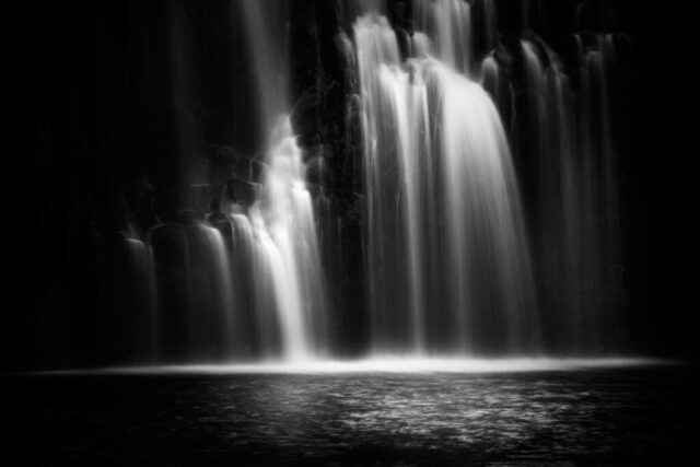 Minimalist Cascade Black and white fine art photograph of McCloud Middle Falls in Northern California, captured in a minimalistic, low-key style. The image isolates a section of the cascade, where soft, silky water flows gracefully over dark volcanic rock, creating a serene and ethereal mood. The long exposure smooths the water into luminous ribbons of li