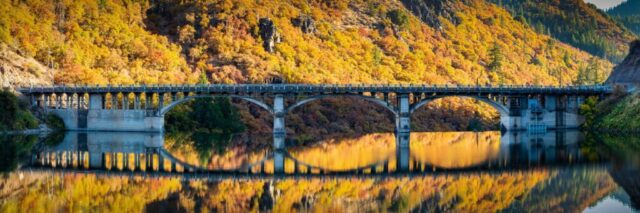 Pit 3 Autumn Panorama A panoramic view of the Pit 3 Bridge spanning the dam at Lake Britton in Shasta County, California. The bridge arches gracefully over calm, reflective water surrounded by steep, autumn-colored hillsides. The lake’s smooth surface creates a mirror-like reflection that resembles an infinity-edge pool against the golden foliage.