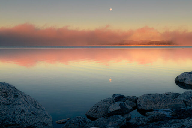 Serene winter morning at Eagle Lake in Northern California, where soft fog drifts across still waters beneath a setting nearly full moon. The rising sun bathes the mist in warm peach and gold hues, reflecting beautifully across the glassy surface and smooth granite shoreline. Subtle mountain silhouettes frame the horizon, adding depth to this tranquil lakeside scene. Captured on a crisp December dawn, this fine art landscape photograph embodies calm, solitude, and the fleeting beauty of light meeting fog over one of California’s most peaceful high-country lakes.