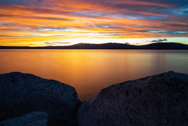 Golden sunset landscape photograph taken from the Lassen County Youth Camp at Eagle Lake, California, where the calm water glows beneath a fiery winter sky. Long exposure smooths the lake’s surface into liquid gold, reflecting brilliant bands of orange, yellow, and indigo as the sun dips behind the distant Sierra Nevada foothills. Granite rocks in the foreground anchor the serene composition, adding texture and contrast to the soft expanse of sky and water. This fine art image captures the tranquil beauty of dusk at Eagle Lake—a quiet moment of stillness and color in Northern California’s high mountain country.