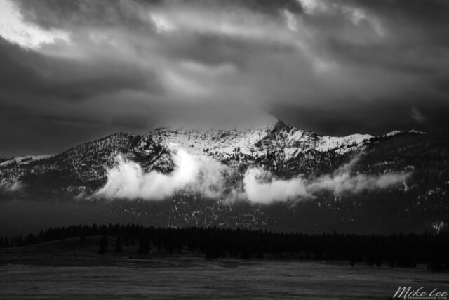 Thompson Peak, straddling the Lassen and Plumas County line in Northern California, rises dramatically in this black and white landscape photograph titled “Thompson Peak Drama.” Captured on a cold January morning in 2021, the image shows the rugged, snow-covered summit shrouded in fast-moving storm clouds as the low winter sun struggles to break through. Wisps of mist cling to the ridgelines, creating the illusion of steam rising from the mountain’s flanks. The dark forested foothills contrast sharply against the glowing snowfields and heavy sky, emphasizing the wild, untamed mood of the Sierra Nevada. This fine art photograph captures the fleeting power and mystery of Thompson Peak’s stormy beauty.