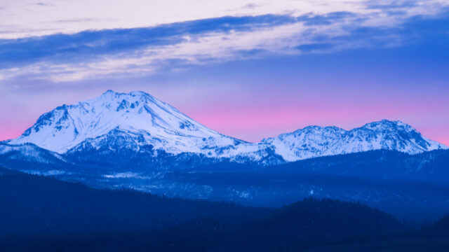 Twilight Majesty Color photograph of Mount Lassen and the Chaos Crags viewed from the Hat Creek Rim in Lassen Volcanic National Park, California, during twilight. The snow-covered volcanic peaks glow softly in the fading light, with cool blue tones blending into a pastel pink and purple horizon. Layers of dark forested ridges stretch toward the mountains, creating depth and serenity. The sky’s gentle gradient and the mountain’s crisp silhouette capture the peaceful transition from golden hour to blue hour on a winter evening.