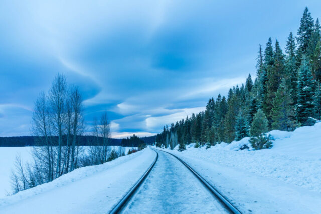Winter scene featuring BNSF railroad tracks curving through a snow-covered landscape near Long Lake in Lassen County, California. The tracks cut a dark line through the bright snow as lenticular clouds form dramatic, layered shapes across the cold blue sky. Evergreen trees border the right side of the frame while bare aspens rise on the left, framing a tranquil yet moody alpine view.