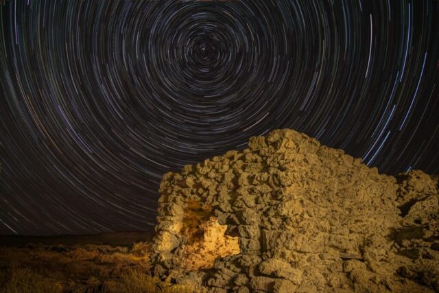 Wheel in the Sky Keeps Burning Long-exposure night photograph titled “Wheel in the Sky Keeps Burning,” featuring the historic Amedee Lime Kiln in Lassen County, California, beneath a mesmerizing swirl of star trails. The image captures approximately 75 minutes of Earth’s rotation centered on Polaris, the North Star, creating a perfect celestial spiral above the rugged stone ruins. Shot deep into astronomical darkness on a moonless night, the scene combines stationary low-level lighting and delicate “light painting” to highlight the kiln’s weathered texture. Once active in the late 19th and early 20th centuries near the vanished town of Amedee, this relic now stands as a haunting monument to a bygone industrial dream — illuminated under the eternal motion of the northern sky.