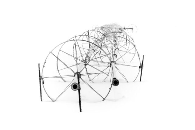 High-key black and white photograph of two irrigation wheel lines standing idle in a snow-covered field in Lassen County, California. The repeating circular forms of the metal wheels and pipes create a rhythmic pattern that fades softly into the white background. The minimalistic composition emphasizes geometry, symmetry, and contrast, evoking a quiet sense of stillness in the frozen winter landscape.