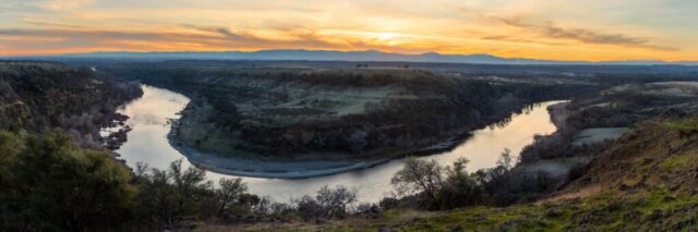 Expansive panoramic landscape photograph of the Sacramento Riverbend in Tehama County, California, showing the dramatic curve of the river winding through rolling green hills under a glowing late winter sunset. Captured from a high vantage point, the 3-to-1 stitched image reveals the tranquil beauty of the Sacramento River as it reflects warm golden light amid the soft gradients of evening sky. Oak-dotted slopes and rugged bluffs frame the serpentine waterway, while distant mountain silhouettes fade into the horizon. This fine art image captures the serene, timeless character of Northern California’s largest river and the natural splendor of the Sacramento Riverbend area.