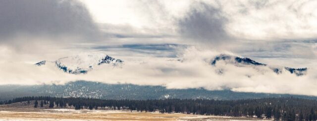 Panoramic view of Thompson Peak on the Lassen and Plumas County line in Northern California, captured from Bass Hill near Highway 395. Snow blankets the mountain slopes as low clouds and fog swirl around the peaks beneath a stormy February sky. Below, a vast golden pasture stretches across the foreground, part of a working cattle ranch framed by dense evergreen forest. The dramatic light and shifting weather reveal the rugged beauty of this high Sierra landscape.