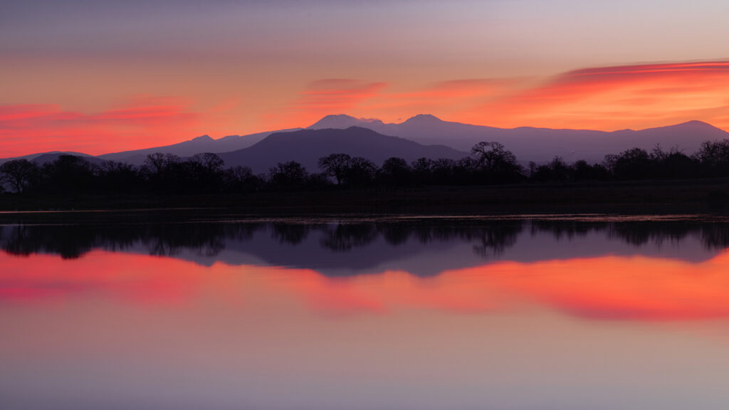 Peaceful dawn landscape photograph captured at Hog Lake in the Sacramento Riverbend area near Red Bluff, California. The image showcases the tranquil stillness of the lake as it mirrors the vivid pink, orange, and violet tones of sunrise spreading across the sky. Silhouetted trees line the far shore, while soft atmospheric haze layers the distant mountains, revealing the distinct forms of Mount Lassen and Chaos Crags in the background. The glasslike reflection and pastel gradients create a sense of calm and balance, capturing the quiet beauty of a Northern California morning at the edge of Lassen Volcanic country.