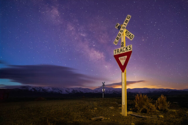 Remote Crossing Nightscape photograph titled “Remote Crossing,” captured in the vast high desert of northwestern Nevada. The image portrays a solitary railroad crossing sign standing beneath the fading night sky as the first light of dawn begins to wash over the landscape. Wisps of cloud reflect the distant glow of Reno, adding soft lavender and golden tones to the horizon. Above, the galactic core of the Milky Way still shines brightly, marking the transition from astro darkness to civil twilight. The scene — both peaceful and desolate — captures the quiet poetry of isolation, where manmade relics meet the boundless sky in a fleeting moment between night and day.