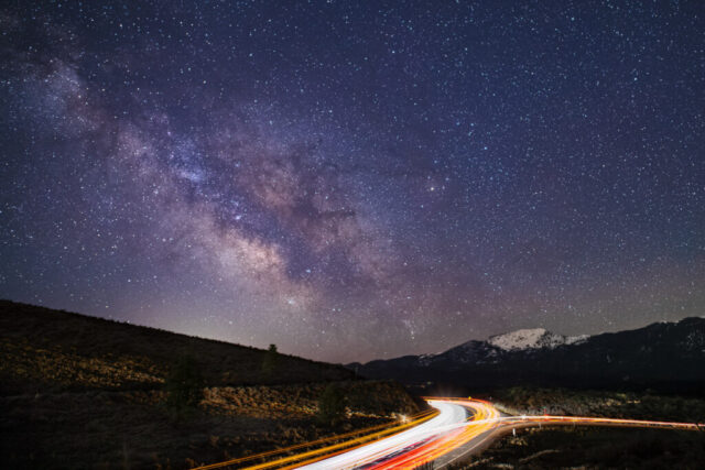 Commute to the Core Nightscape photograph titled “Commute to the Core,” captured along U.S. Highway 395 at Bass Hill in Lassen County, California. The image looks toward the southeast, where the snow-dusted peak of Thompson Mountain rises beneath the dazzling Milky Way core. Long-exposure captures of passing vehicles create glowing red and white light trails that trace the roadway, symbolizing the quiet intersection of human motion and cosmic stillness. Above, countless stars and interstellar clouds stretch across the night sky in vivid detail — the result of multiple stacked exposures blended to reduce digital noise. This piece perfectly balances the earthly and the celestial, offering a serene reminder of our small yet luminous presence in the grand commute of the universe.