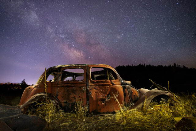 Rusting Under the Stars Nightscape photograph titled “Rusting Under the Stars,” featuring an abandoned vintage car resting in Ranch Park near Susanville, California. The rusted, timeworn vehicle sits quietly in a field of wild grass beneath the brilliant galactic core of the Milky Way. The long-exposure composite blends multiple stacked frames for a clean, noise-free sky and several light-painted exposures of the car, revealing rich texture and color in its corroded metal. Subtle purples and golds illuminate the night horizon while the Milky Way’s glowing band stretches across the sky above the silhouette of distant trees. This evocative image captures the beauty of decay and endurance — a relic of human history set against the timeless expanse of the cosmos.