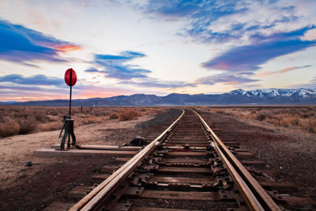 Fine art landscape photograph captured at sunrise on a remote railroad spur in the northwestern Nevada desert, between Pyramid Lake and the California state line. The composition features a steel rail switch in the foreground, its red signal standing out against the warm dawn light as the twin tracks curve gently toward distant mountains beneath a colorful sky. Soft pink, orange, and blue clouds stretch across the horizon, reflecting the first light of day over the high desert. This image blends industrial geometry with natural beauty, symbolizing both direction and solitude in the quiet stillness of a desert morning.