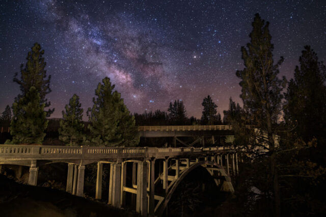 Devils Corral Nightscape Nightscape photograph titled “Devils Corral Nightscape,” featuring the historic Devil’s Corral Bridge and the rebuilt railroad trestle spanning the Susan River near Susanville in Lassen County, California. The image, captured in the early hours of a March morning, showcases the abandoned 1923 concrete arch bridge beneath the luminous galactic core of the Milky Way. The old structure, illuminated through careful light painting, contrasts beautifully with the dark silhouettes of surrounding pines and the softly glowing night sky. Above it all, countless stars shimmer across a magenta-hued horizon, while the Milky Way’s dense band of starlight arcs gracefully overhead. This long-exposure composition blends multiple frames for a noise-free, richly detailed view — a hauntingly beautiful tribute to the region’s transportation history and the quiet endurance of manmade relics beneath the timeless sky.
