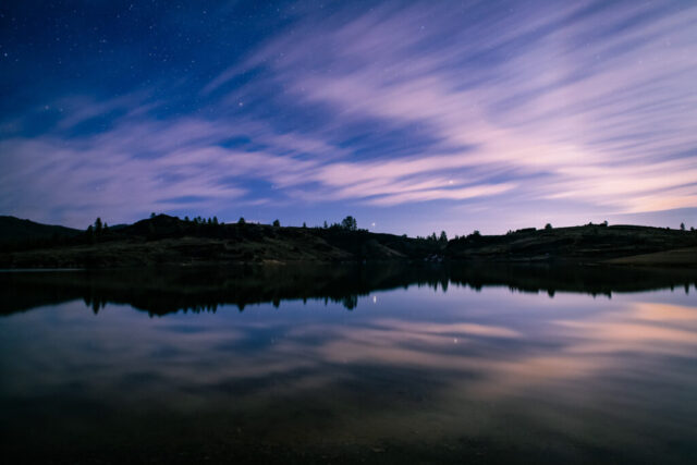 Frenchman Lake Nightscape Nightscape photograph of Frenchman Lake in Plumas County, California, captured under a softly clouded sky just after twilight. The long-exposure image shows streaks of lavender and blue clouds drifting across the heavens, faint stars twinkling through the gaps, and their reflections mirrored perfectly on the calm surface of the lake. The dark silhouette of rolling hills and scattered pines frames the horizon, while a single bright celestial object — likely a planet — glows near the center, adding a point of brilliance to the tranquil scene. This serene composition captures the quiet beauty of the Northern Sierra Nevada night, where light, motion, and reflection blend into a painterly display of peace and color.