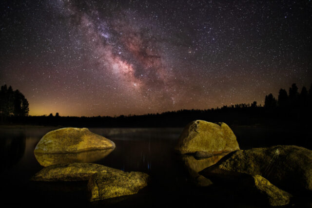Antelope Lake Nightscape Nightscape photograph titled “Antelope Lake Nightscape,” featuring the Milky Way’s galactic core rising above the still waters of Antelope Lake in Plumas County, California. Captured during the pre-dawn hours of a cold spring morning, the image reveals a few massive boulders along the shoreline, gently illuminated through light painting. Faint fog begins to form over the water as the air meets the dew point, subtly diffusing reflections of stars shimmering between the rocks. The galactic core glows in vivid tones of gold, magenta, and violet against the dark silhouette of the pines, while a soft horizon glow from the towns of Portola and Quincy adds warmth to the scene. This long-exposure, multi-frame composition captures the quiet, fleeting beauty of starlight, fog, and frozen air in the Sierra Nevada high country.