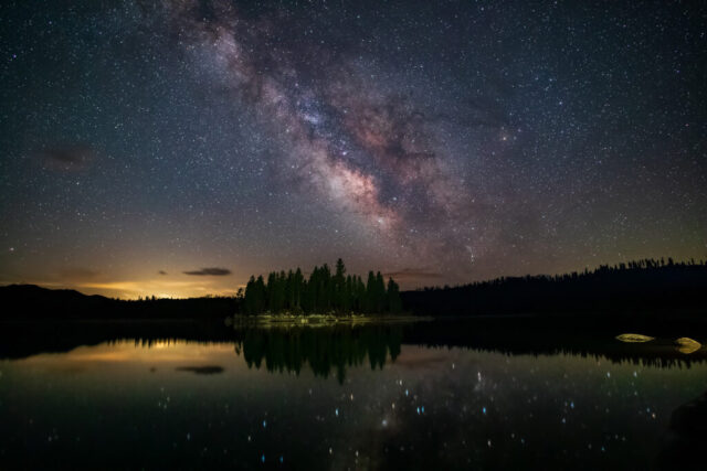 Jewels on the Water Nightscape photograph titled “Jewels on the Water,” featuring the Milky Way’s galactic core rising over Antelope Lake in Plumas County, California. Captured on a crystal-clear April morning, the image reveals countless stars reflected on the calm surface of the lake, surrounding a small pine-covered island near the Lost Cove Boat Ramp. The faint glow of Portola, California, softly illuminates the horizon to the left, while subtle wisps of clouds add depth to the night sky. The long-exposure composition, created by stacking multiple frames for clarity and noise reduction, showcases the Milky Way’s brilliant core in hues of violet, rose, and gold — mirrored like scattered gemstones upon the tranquil Sierra Nevada waters.