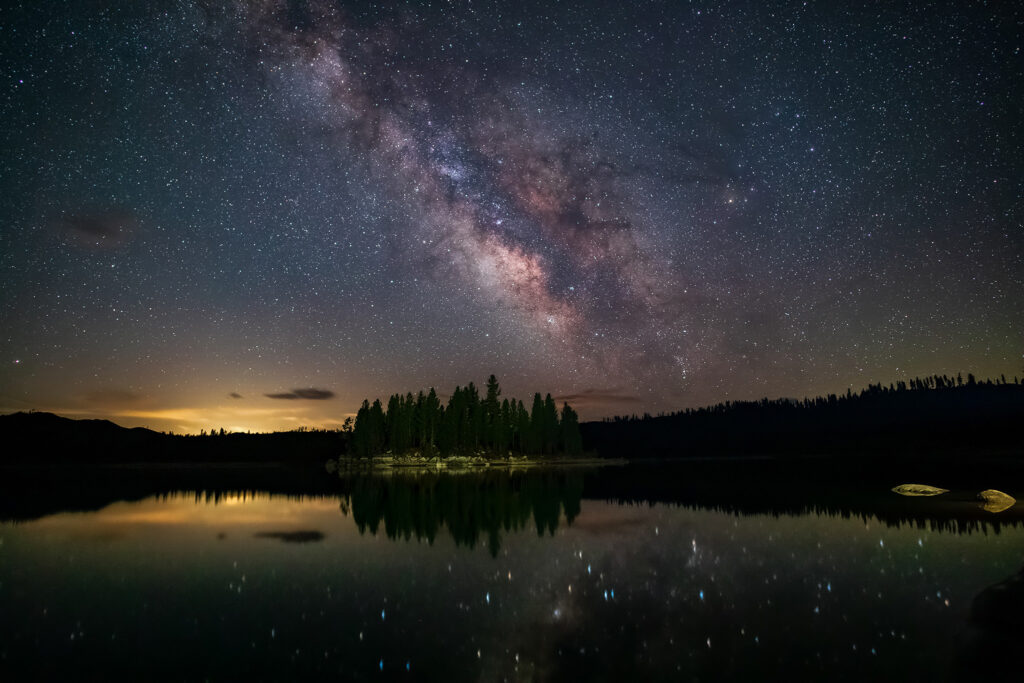 Nightscape photograph titled “Jewels on the Water,” featuring the Milky Way’s galactic core rising over Antelope Lake in Plumas County, California. Captured on a crystal-clear April morning, the image reveals countless stars reflected on the calm surface of the lake, surrounding a small pine-covered island near the Lost Cove Boat Ramp. The faint glow of Portola, California, softly illuminates the horizon to the left, while subtle wisps of clouds add depth to the night sky. The long-exposure composition, created by stacking multiple frames for clarity and noise reduction, showcases the Milky Way’s brilliant core in hues of violet, rose, and gold — mirrored like scattered gemstones upon the tranquil Sierra Nevada waters.