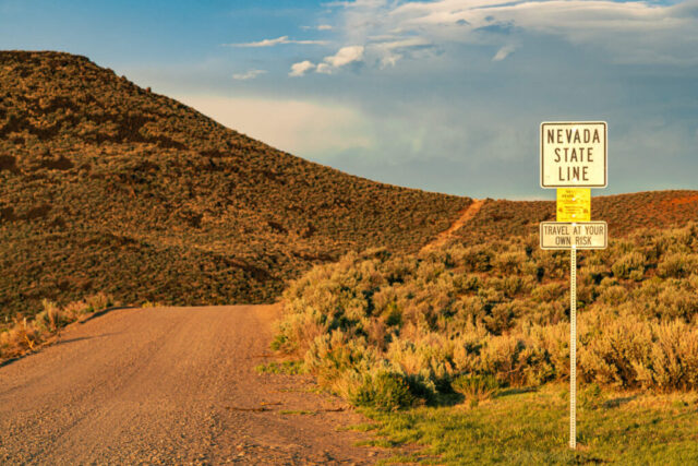 A dusty stretch of Smoke Creek Road disappears into the rugged hills beneath warm evening light, marking the remote Nevada state line. The weathered roadside sign stands as both a boundary and a warning—“Travel at Your Own Risk”—perfectly capturing the wild, unpolished character of the high desert frontier. Sagebrush and golden hills surround the dirt track, glowing under late spring sun in this timeless image of the American West. A compelling photograph that embodies solitude, open-road adventure, and the untamed spirit of the California–Nevada borderlands.