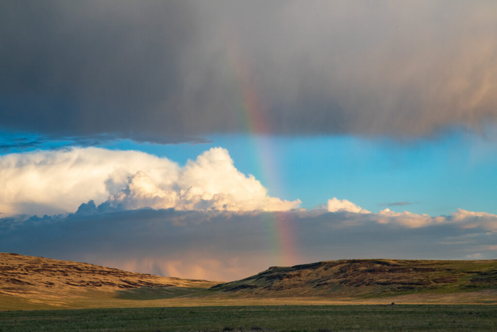 A faint rainbow arcs across dramatic spring storm clouds above the rugged hills of the Smoke Creek Desert in Lassen County, California. Golden light warms the arid landscape below as layers of blue sky, white cumulus, and gray rainclouds blend in a vivid high-desert scene.