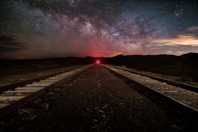 Gallactic Railroad Nightscape photograph titled “Galactic Railroad,” featuring the Burlington Northern Santa Fe railroad spur beneath the glowing Milky Way core at Flannigan in northwestern Nevada. Captured on a hazy late-spring evening, this long-exposure image combines stacked sky frames for reduced digital noise with light-painted foreground exposures of the tracks. The brilliant red signal light at the end of the rail line casts an ethereal glow that reflects off the thin haze, adding a dramatic contrast against the dark mountains and starlit sky. The Milky Way arcs majestically above in hues of violet, rose, and gold, while faint orange illumination from Reno lights the clouds on the horizon. This photograph evokes both the industrial legacy of Nevada’s railroads and the timeless beauty of the cosmos — where human craftsmanship meets the infinite sweep of the universe.