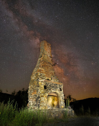 Cosmic Fireplace Nightscape photograph of the historic roadside fireplace and chimney along California State Highway 36 in Lassen County, California, captured beneath the brilliant galactic core of the Milky Way. The towering stone and brick structure, all that remains of a 1930s bar, restaurant, and gas station once operated by Bill and Jeanette Tunison, glows warmly through careful light painting. Behind it, the dense star fields and golden hues of the Milky Way stretch across the night sky, adding a sense of cosmic depth to the scene. This site, now maintained by the Chimney Fund—a local philanthropic group that decorates it for holidays—stands as both a relic of regional history and a striking visual anchor against the vast celestial backdrop. Created using multiple stacked sky exposures and blended flashlight-lit frames, the image highlights the intersection of human legacy, rural history, and the timeless wonder of the night sky.