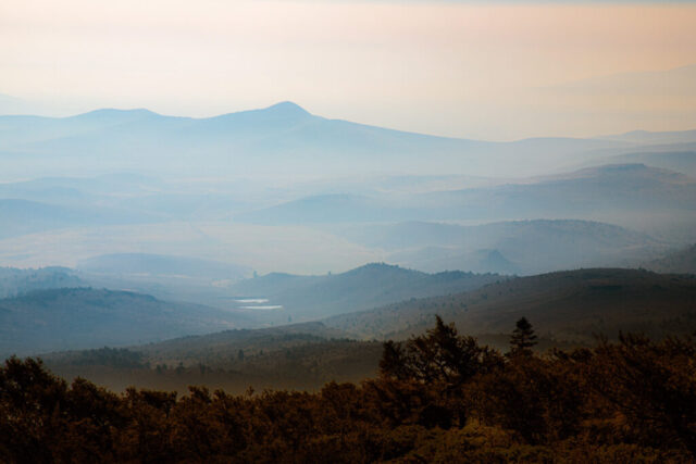 Photograph taken from the summit of Fredonyer Peak in Lassen County, California, showing a layered, smoky mountain vista at sunrise during the 2021 Dixie Fire. The scene reveals ridges and valleys fading into the distance under a soft orange and blue haze caused by atmospheric inversion, which trapped wildfire smoke in the lower elevations. Taken from the 7,949-foot peak near the U.S. Forest Service lookout managed by the Bureau of Land Management, this image captures both the tranquil beauty and somber reality of Northern California’s largest recorded single wildfire. A compelling fine art landscape photograph emphasizing mood, depth, and the natural resilience of the Sierra Nevada region." />