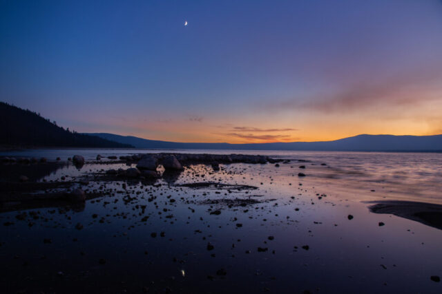 Tranquil twilight landscape photograph taken at Miner’s Point on Eagle Lake in Lassen County, California, during the peaceful blue hour of a mid-September evening. A slender crescent moon hangs above the fading orange and violet horizon, its reflection shimmering faintly in the shallow water among scattered shoreline rocks. Gentle ripples and soft light evoke stillness beneath the vast mountain silhouettes that frame the high-altitude lake. This fine art image captures the quiet solitude and natural beauty of Eagle Lake—one of California’s largest natural lakes—where calm water, twilight color, and moonlight meet in serene harmony.