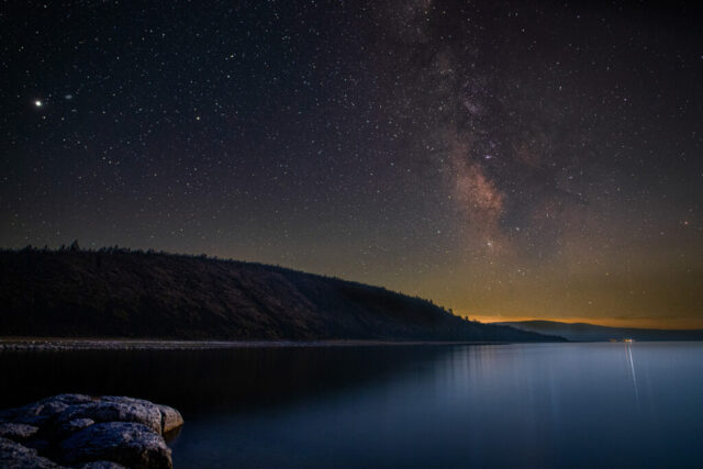 Miner's Point Starscape Nightscape photograph titled “Miner’s Point Starscape,” captured at Miner’s Point on Eagle Lake in Lassen County, California. The image showcases the Milky Way rising nearly vertical in the late-summer sky, its galactic core glowing in rich tones of magenta, gold, and violet above the dark shoreline. A 25% crescent moon, just out of frame to the west, casts gentle light across the lake’s calm surface, revealing subtle textures in the distant hills. Reflections from Gallatin Marina glimmer faintly on the water, while the planet Saturn shines brightly near the upper left of the frame. Created by stacking multiple sky exposures for reduced noise and blending them with a single moonlit foreground frame, this long-exposure composition captures the serene beauty and quiet scale of Eagle Lake — a high-elevation wilderness haven renowned for its rainbow trout and timeles