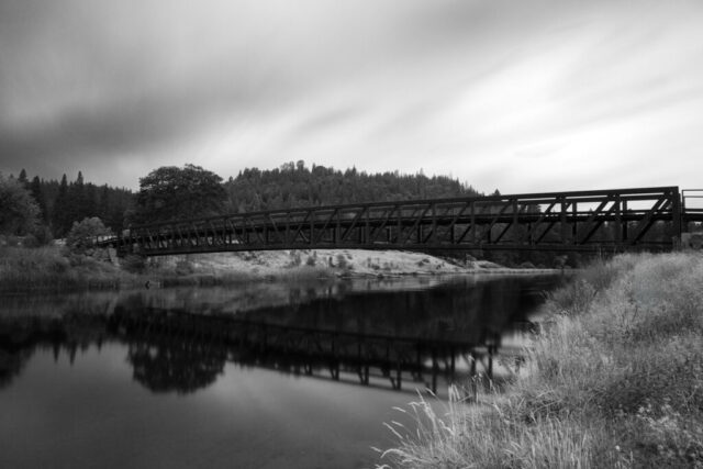 Rustic Bridge Over Hat Creek - Monochrome Monochrome long-exposure photograph of the bridge spanning Hat Creek near the Hat #2 Powerhouse in Cassel, Shasta County, California. Captured on a rainy September evening, this image transforms a missed sunset into a study of tone and texture. The bridge’s angular steel structure contrasts beautifully with the softened reflections and streaking clouds, creating an atmosphere both serene and dramatic. A timeless black and white rendering that highlights the harmony between man-made design and natural beauty—perfect for admirers of fine art landscapes, architectural photography, and the quiet moods of the Northern California wilderness.