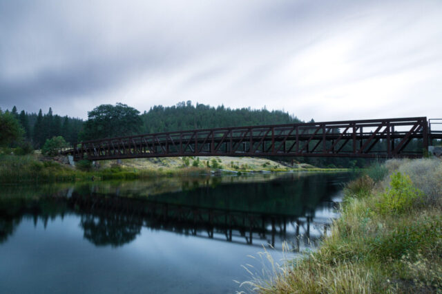 Rustic Bridge Over Hat Creek Long-exposure photograph of the rustic bridge spanning Hat Creek near the Hat #2 Powerhouse in Cassel, Shasta County, California. Captured on a moody September evening as rain began to fall, the image features the dark steel bridge reflected in the still creek waters beneath a textured, cloud-streaked sky. The surrounding pines and golden grasses frame the tranquil scene, highlighting the serene character of this blue-ribbon trout stream. A contemplative landscape that evokes quiet solitude—perfect for lovers of Northern California scenery, fishing country, and atmospheric fine art photography.