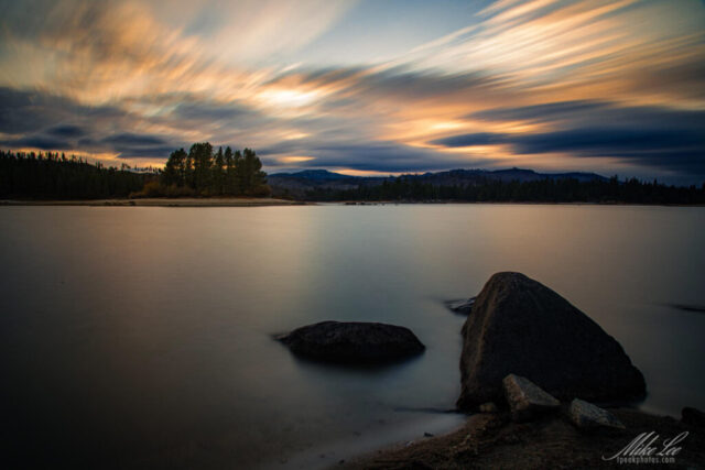 Dramatic long exposure photograph of Antelope Lake in Plumas County, California, captured from Lunker Landing as a small storm approached at sunset. The smooth, glassy water contrasts beautifully with streaking clouds that radiate warm tones of gold and orange against the darkening sky. In the foreground, large rocks anchor the tranquil lake scene while a small island of pines sits peacefully in the distance. Taken shortly after the Dixie Fire, this image conveys renewal, serenity, and the quiet power of nature in motion—a stunning fine art landscape evoking calm before the storm.
