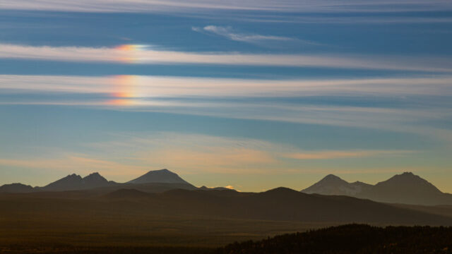 A parhelion, or “sun dog,” glows above the Three Sisters peaks near Bend, Oregon, with soft, colorful light refracting through thin cirrus clouds. The volcanic mountains rise in silhouette against a tranquil blue and gold sky, viewed from the top of Pilot Butte overlooking the high desert landscape below.