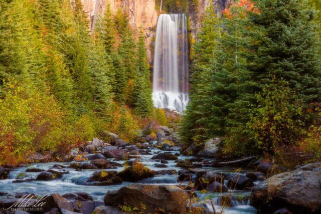 Tumalo Falls Autumn Dawn Photograph of Tumalo Falls near Bend, Oregon, a magnificent 97-foot waterfall on Tumalo Creek in the Deschutes National Forest. Captured on an October morning shortly after sunrise, the scene glows with autumn color as golden and amber foliage lines the stream below the towering cascade. The water flows gracefully through a lush canyon framed by evergreens and volcanic rock, embodying the wild beauty of Central Oregon. Processed with a soft painterly touch, this fine art image evokes serenity, renewal, and the timeless artistry of nature’s landscapes in the Pacific Northwest.