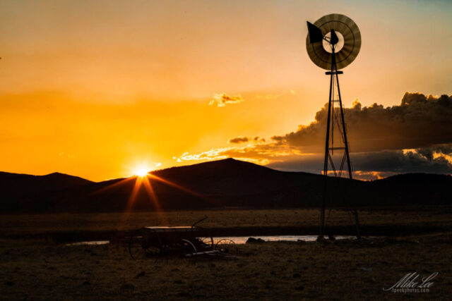 Aermotor and the Setting Sun A stunning sunset photograph of a classic Aermotor windmill standing in a quiet pasture south of Madeline in Lassen County, California. The setting sun dips behind distant hills, casting golden light across the sky and reflecting off the still water below. Silhouetted against the brilliant orange and amber glow, the old wind pump rises proudly, its blades frozen in the calm evening air. This peaceful scene captures the enduring spirit of rural California ranch life—simple, timeless, and beautifully illuminated by the last light of day.