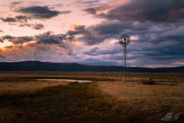 Aermotor Evening Shift A dramatic evening landscape featuring a classic Aermotor windmill standing tall in a wide-open pasture south of Madeline in Lassen County, California. The scene is bathed in the rich, moody light of sunset as deep clouds swirl across the sky in shades of violet, orange, and gold. The weathered wind pump, silhouetted against the colorful horizon, embodies the quiet endurance of rural life—steadily drawing water for cattle beneath the vast high-desert sky. This image captures the timeless beauty and resilience of working ranchland in northern California’s Great Basin country.