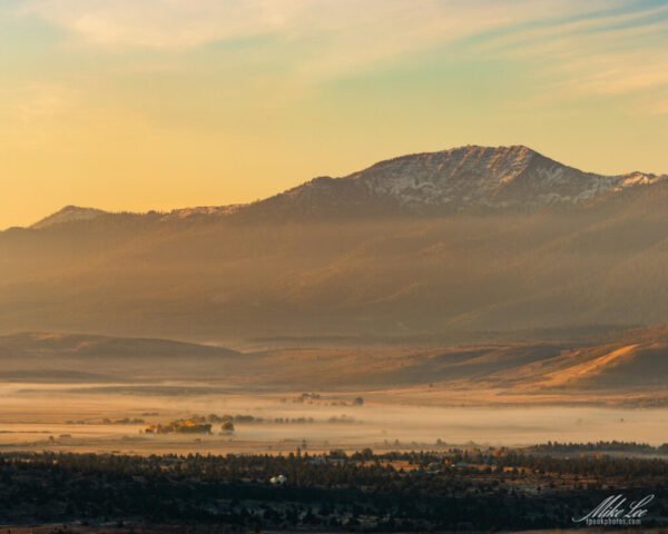 Golden sunrise over Honey Lake Valley in Lassen County, California, with Thompson Peak rising in the background. A thin layer of fog drifts through the valley below the rugged, snow-dusted mountain as warm early morning light bathes the landscape in amber tones. Wisps of mist and soft shadows add depth to the foothills and sage-covered slopes, capturing the tranquil beauty of a foggy autumn morning in the high desert.
