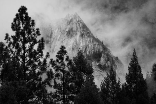 Black and white landscape photograph of the dramatic granite spires of Castle Crags State Park near Castella, in Northern California’s Shasta-Trinity National Forest. The image captures the raw, moody atmosphere of a stormy day, with mist and rain drifting through the scene as jagged peaks rise sharply behind silhouetted pine trees in the foreground. The interplay of fog, shadow, and texture emphasizes the ancient, glacially carved rock faces and the sense of solitude in this rugged wilderness. The monochrome treatment enhances the stark drama and timeless beauty of the crags, evoking the spirit of exploration and the wild, untamed landscapes of the Shasta Cascade region.