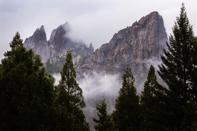 A moody landscape photograph of Castle Crags in Shasta County, California, capturing jagged granite spires rising through low-hanging mist and drifting storm clouds. Taken from Exit 723 along Interstate 5 near Dunsmuir, this dramatic scene highlights the rugged texture and vertical drama of the Crags, remnants of ancient glaciation that carved the region’s peaks. The composition frames the gray stone towers behind dark evergreen pines, their silhouettes enhanced by soft diffused light filtering through the passing storm. The image conveys a sense of mystery, solitude, and natural grandeur—an evocative portrait of Northern California’s wild mountain terrain perfect for outdoor, fine art, and nature photography enthusiasts.